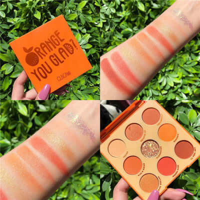 Swatches of the Orange You Glad eyeshadow palette on a wrist surrounded by green foliage.